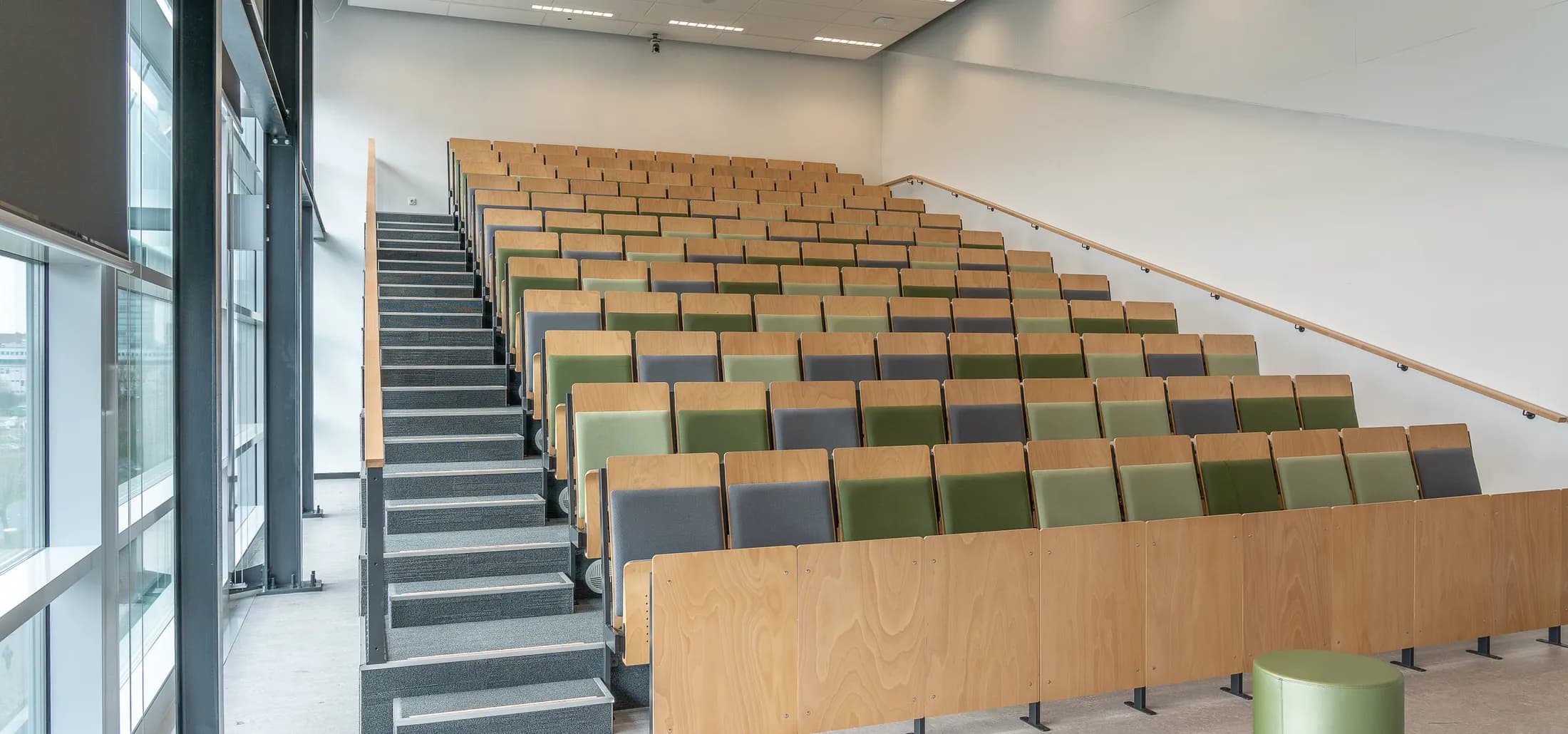 An empty auditorium of tiered wooden seats in green and grey, daylight through a glass facade at the left