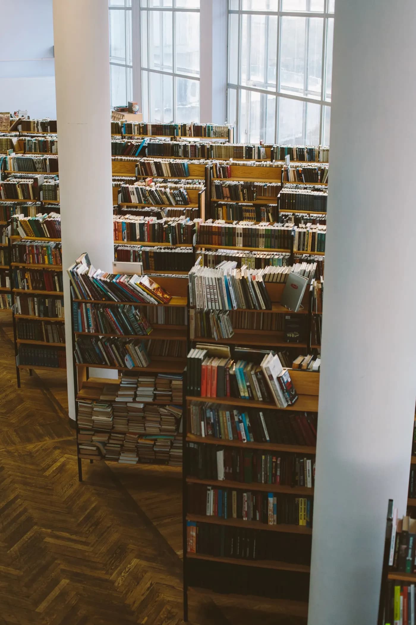 Der Lesesaal einer Bibliothek zwischen zwei weißen Säulen, Holzregale voller Bücher, hohe Fenster und Parkettboden, niemand zu sehen
