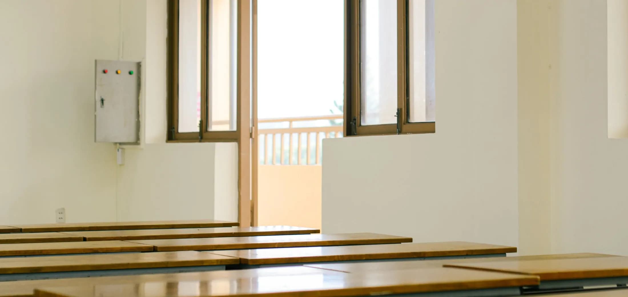 Empty rows of long pale desks in a teaching room, tall windows standing open to the daylight.
