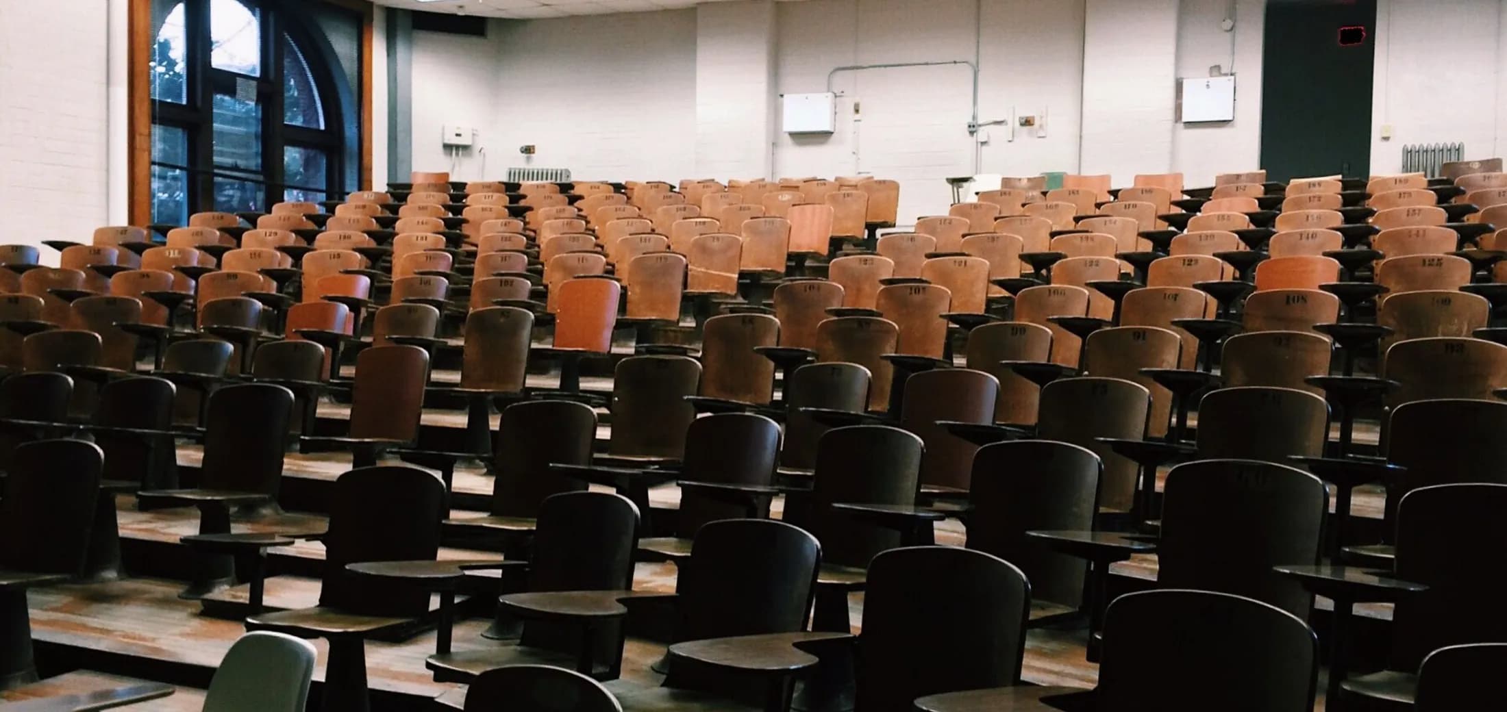A large lecture hall, rows of empty tip-up seats with writing tablets, daylight from tall windows at the back