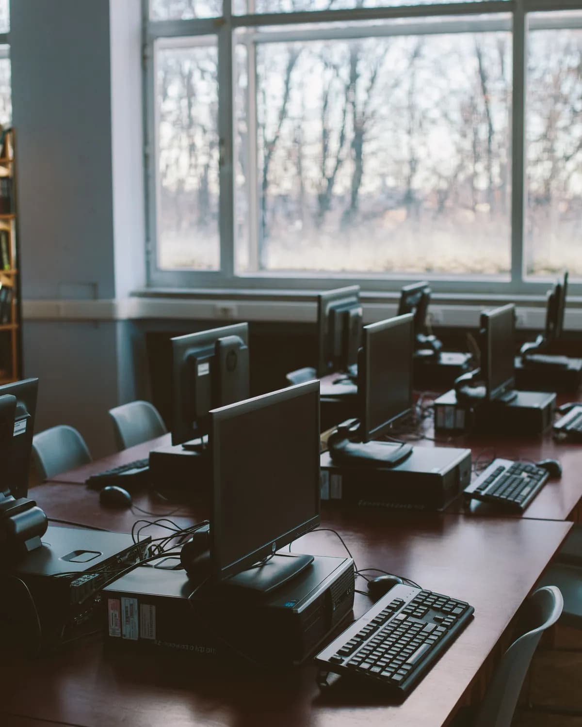 An empty school computer room, screens and keyboards on long wooden desks, winter light through the windows behind
